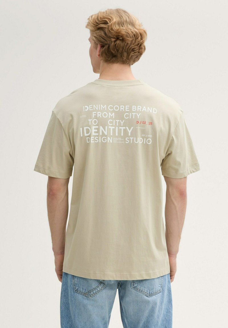 Beige katoenen T-shirt met korte mouwen, met witte tekst op de achterkant: branding en ontwerpdetails in verschillende lettertypen. Spijkerbroek zichtbaar.