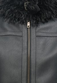 Svart läderjacka med en strukturerad yta, med en tjock svart pälskrage, exponerad metalldragkedja och två horisontella bröstfickor.