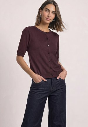 Femme aux cheveux mi-longs portant des boucles d’oreilles créoles, un cardigan bordeaux à manches courtes boutonné et un jean large foncé, les mains dans les poches.