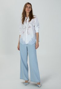 White button-up shirt with intricate blue embroidery at the hem, paired with light blue wide-leg jeans and shiny silver shoes.