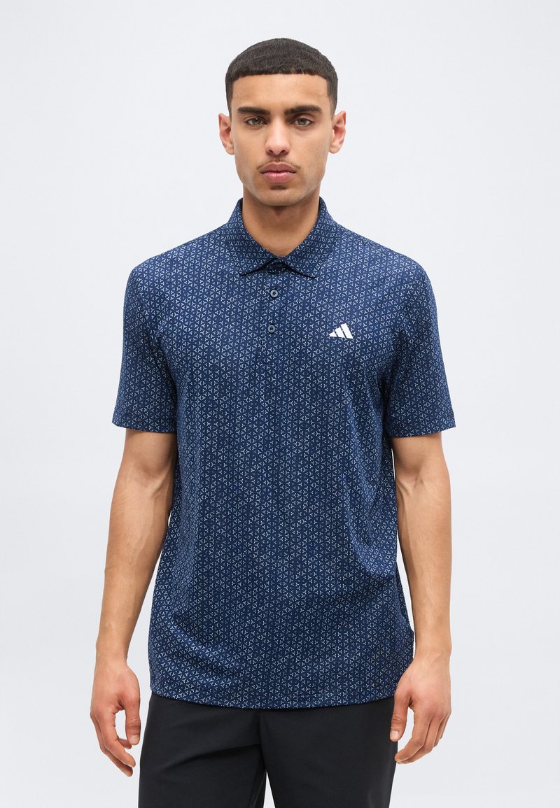 Νέος άνδρας φορώντας μπλε σκούρο πόλο με κοντά μανίκια και σχέδιο, με λευκό λογότυπο στο στήθος, στέκεται μπροστά σε απλό ανοιχτό σκηνικό.