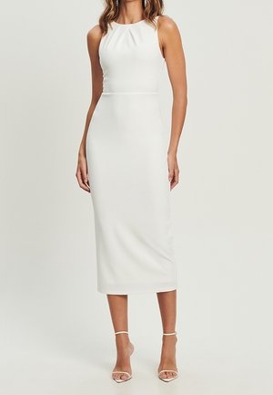 Robe midi blanche sans manches, cintrée, avec un col montant, portée par une personne debout, accompagnée de sandales blanches à talons hauts et à lanières.