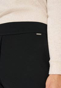 Svarta stretchbyxor med en slät textur, med en åtsittande midja och en liten metall-logotypdetalj. Intill en ljus tröja.