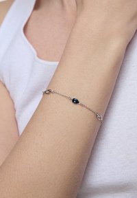 Srebrna bransoletka łańcuszkowa z akcentem w postaci niebieskiego kamienia w kształcie łzy, delikatne ogniwa i pewny zamek, noszona na nadgarstku.