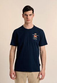 Camiseta de algodón azul marino con cuello redondo, que presenta logotipos bordados en rojo, amarillo y blanco. Mangas cortas, corte relajado.