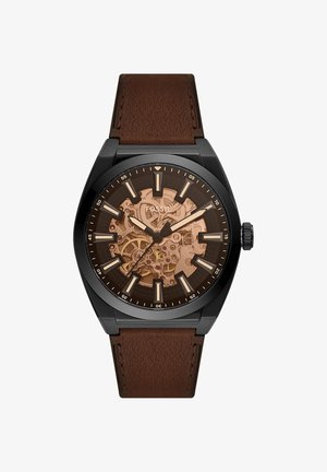 Orologio da polso da uomo con cassa nera, cinturino in pelle marrone, ingranaggi meccanici in bronzo esposti e indici e lancette luminescenti.