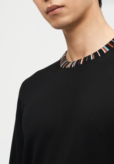 Homme portant un pull noir avec un col rayé multicolore, visible des épaules au menton sur un fond uni.