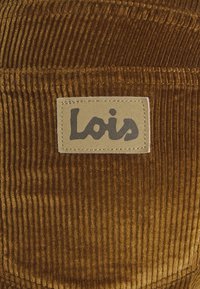Tissu en velours côtelé marron avec une étiquette beige cousue portant le nom de la marque "Lois" en lettres foncées.