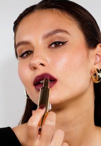 Pomadka w złotej tubce trzymana blisko ciemnowiśniowych ust. Tubka ma fakturowaną powierzchnię i jest w użyciu. Kolczyki to okrągłe złote kółka.