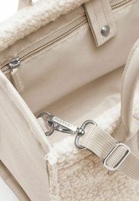 Borsa di tessuto beige con un morbido interno in pile testurizzato, dotata di una tasca con zip, accessori metallici e una tracolla staccabile.