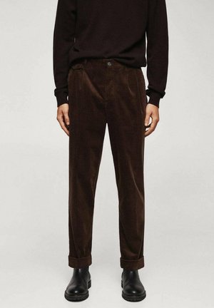 Person, die dunkelbraune Cordhose mit Umschlag, einen schwarzen Pullover und schwarze Lederstiefel trägt, steht vor einem neutralen Hintergrund.