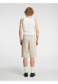 Beige cargoshorts med to baklommer, kombinert med en hvit tanktop. Modellen har på seg hvite sokker og svarte sko. Teksturen ser glatt ut.