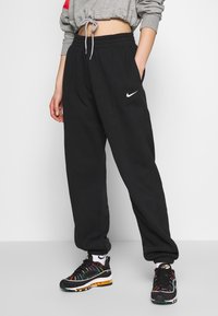 Černé joggingové kalhoty Nike s elastickým pasem, postranními kapsami a bílým logem na levém boku, doplněné barevnými sportovními botami.