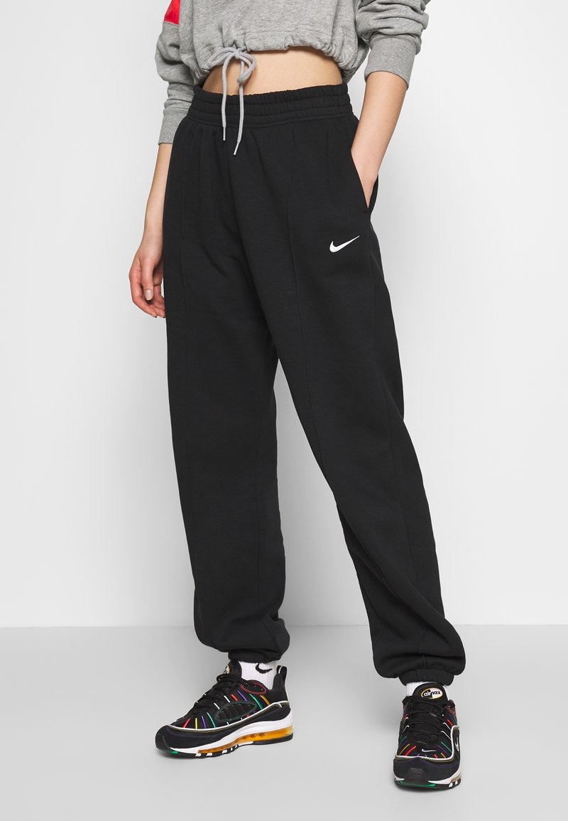 Černé joggingové kalhoty Nike s elastickým pasem, postranními kapsami a bílým logem na levém boku, doplněné barevnými sportovními botami.