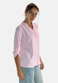 Jasnoróżowa, długorękawowa koszula zapinana na guziki z podwiniętymi rękawami, kołnierzykiem henley i delikatną fakturą. Noszona z jasnoniebieskimi dżinsami.