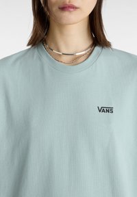 Světle modré tričko s krátkým rukávem vyrobené z bavlny. Na přední části v levém hrudním místě se nachází černé logo Vans a má design s kulatým výstřihem.