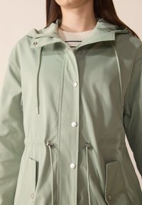 Jachetă impermeabilă de culoare verde deschis, cu glugă, nasturi tip snappy, șnururi la talie și două buzunare frontale. Textură netedă.