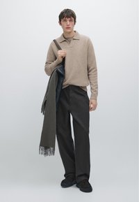 Beige stickad polo-tröja, mörka byxor, håller en grön scarf. Enkel design, mjuk textur, med en avslappnad passform och minimalistiska detaljer.