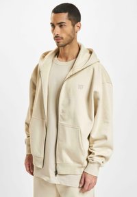 Cremefarbener Zip-Hoodie mit verstellbarem Kordelzug an der Kapuze, großen Fronttaschen und weichem Material. Kombiniert mit einem passenden cremefarbenen T-Shirt darunter.