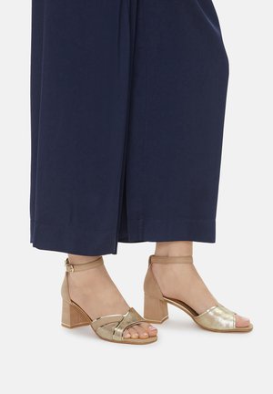 Beige Sandalen mit Absatz und Knöchelriemen, die eine Kombination aus glatten und strukturierten Materialien sowie ein Kreuzband-Design aufweisen.