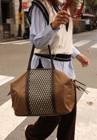 Brown tote bag with woven black geometric pattern, leather accents, sturdy handles, and zippers on the sides, being held by a person.