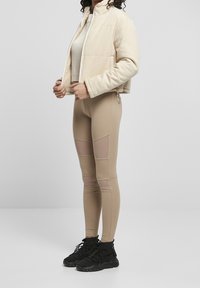 Beige kordjacka i cropped modell över en ljus stickad topp, i kombination med hudfärgade leggings med nät-inserts. Svarta hög-top sneakers fullända looken.