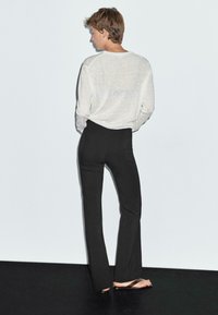 Hellgraues, langärmliges Hemd aus weichem Material, kombiniert mit schwarzen, eng anliegenden Hosen. Das Modell trägt braune Flip-Flops, Ansicht von hinten.