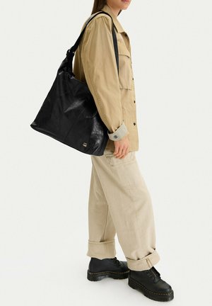 Persona con giacca e pantaloni beige che tiene una grande borsa a tracolla in pelle nera, indossa scarpe platform nere, rivolta verso destra.