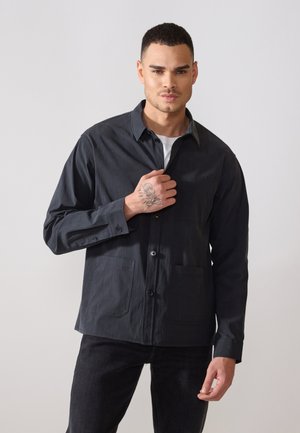 Dark-colored shirt made of textured fabric, featuring a button-up front, a collar, and two front pockets. Casual fit with long sleeves.