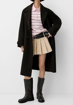 Vrouw draagt een lange zwarte jas, een roze gestreept poloshirt, een beige geplooide rok, een zwarte dubbele riem en zwarte laarzen tot halverwege de kuit tegen een witte achtergrond.