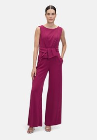 Fuchsia ärmlös jumpsuit med figursydd överdel, vida ben och slående rosett vid midjan. Tillverkad av ett mjukt tyg med en modern design.