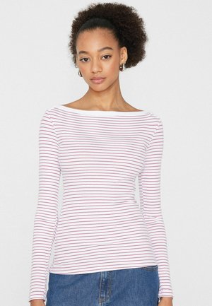 Junge Frau mit natürlichem lockigem Haar trägt ein weißes Langarmshirt mit dünnen roten horizontalen Streifen und blaue Jeans, steht vor einem einfarbigen Hintergrund.