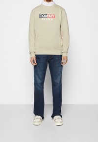 Felpa beige con logo "TOMMY JEANS", abbinata a jeans di denim blu scuro e sneakers color camoscio con motivo mimetico.