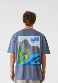 Pelēks T-krekls ar krāsainu ilustrāciju, kas attēlo piekrastes ainavu, un uzrakstu "AMALFI COAST" treknām zilām burtiem. Īsas piedurknes, brīvs piegriezums.
