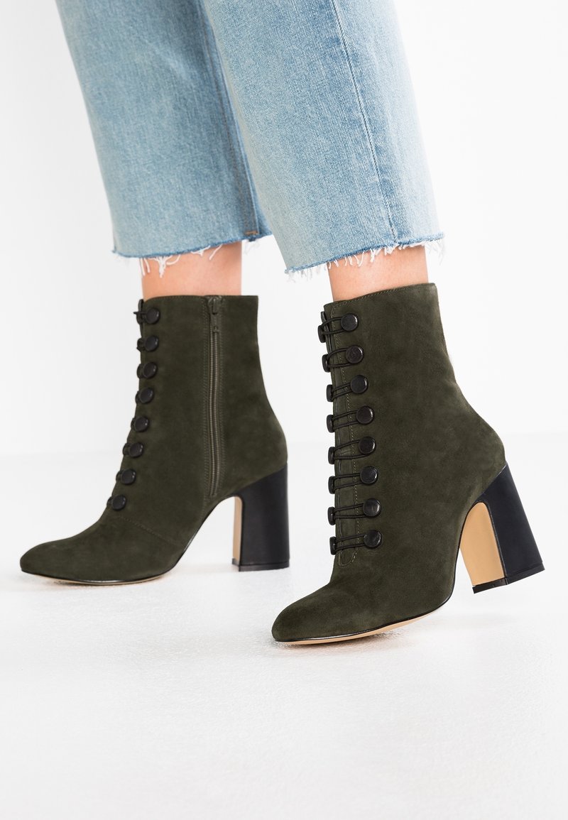 Botas de tobillo de ante verde oliva con puntera puntiaguda, detalles de metal y un robusto tacón negro en bloque. Cuentan con una cremallera lateral y botones decorativos.