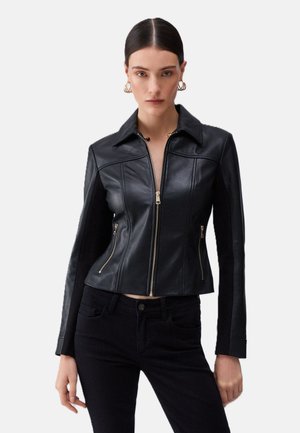 Donna che indossa una giacca di pelle nera aderente con cerniera frontale e tasche con zip, abbinata a pantaloni neri, in piedi davanti a uno sfondo semplice.