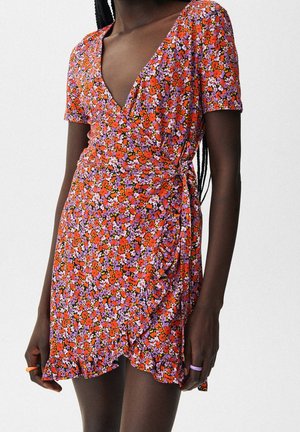 Kobieta ubrana w sukienkę kopertową z krótkim rękawem o czerwono-fioletowo-białym kwiatowym wzorze, z falbaną u dołu i wiązaniem na boku w talii.