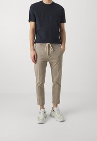 Marineblauwe T-shirt met reliëftekst, gecombineerd met beige jogpants met koord. De schoenen hebben een robuust ontwerp in witte en lichtgele tinten.