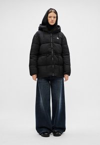 Musta toppatakki, jossa on vetoketjukiinnitys, säädettävä nauhavyötärö ja huppu. Yhdistetty leveälahkeisiin tummansinisiin farkkuihin ja mustiin kenkiin.