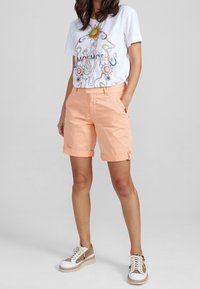Camiseta blanca con un diseño gráfico colorido, combinada con pantalones cortos de durazno claro con puños enrollados. Usada con zapatillas blancas y beige.