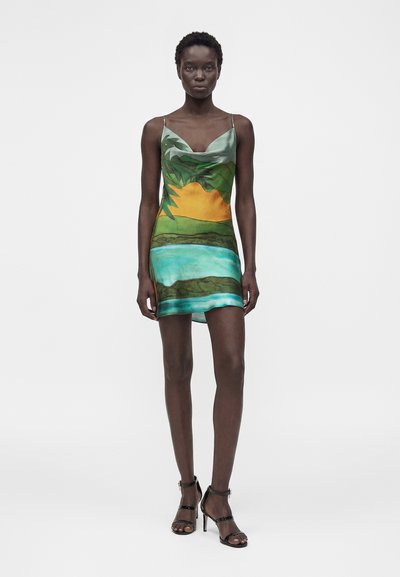 Γυναίκα που φοράει μίνι φόρεμα χωρίς μανίκια με εκτύπωση τροπικού ηλιοβασιλέματος και υδάτινου τοπίου, σε συνδυασμό με μαύρα σανδάλια με ψηλό τακούνι.