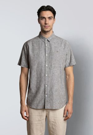 Hombre de pie con una camisa gris de manga corta abotonada y con cuello, y pantalones beige, sobre un fondo liso.