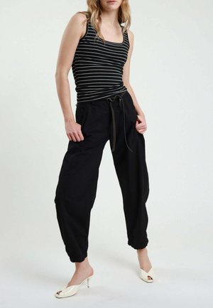 Donna che indossa una canottiera a righe bianche e nere, pantaloni neri larghi con coulisse e tacchi alti bianchi aperti davanti, in piedi con le mani in tasca.