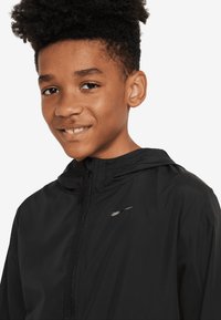 Schwarze Nike Windjacke mit Reißverschluss, Kapuze und Logo auf der Vorderseite. Sie verfügt über eine glatte Textur und ein leichtes Material.