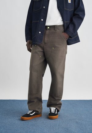 Uomo che indossa pantaloni da lavoro marroni con ginocchia rinforzate, scarpe da ginnastica nere con lacci bianchi, una camicia bianca e una giacca di jeans scura.