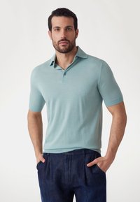 Gaiši zils īsām piedurknēm polo krekls mīkst�ā audumā ar apkakli un divām pogām. Apvienots ar tumši ziliem biksēm ar pogu detaļu.