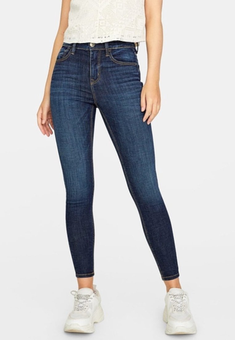 Femme portant un jean skinny taille haute bleu foncé, un haut blanc à motifs et des baskets blanches épaisses, debout sur un fond clair uni.