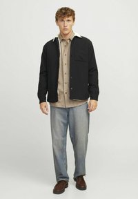 Zwarte button-up jas met een crèmekleurige kraag, gedragen over een beige geruite shirt. Loszittende denim broek en bruine schoenen maken de outfit compleet.