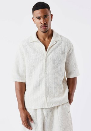 Chemise à manches courtes de couleur crème avec un motif texturé, coupe ample et boutonnière à l'avant. Le col est plat et elle a une esthétique décontractée.