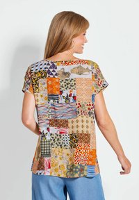 Bluse mit kurzen Ärmeln, die ein Patchwork-Design mit verschiedenen floralen und geometrischen Mustern in Orange, Blau, Gelb und neutralen Tönen zeigt.